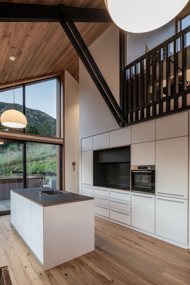 A contemporary open-plan kitchen with a compact island, white handleless cabinetry, dark benchtops, integrated oven, and a dark tiled backsplash. The kitchen has a warm timber ceiling and timber flooring, dramatic black structural beams and mezzanine balustrade, and large floor-to-ceiling windows overlooking a rugged green mountainous landscape. A sculptural pendant light hangs above the island.