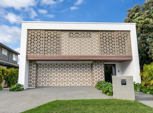 Front exterior view of a two-storey contemporary residence with a flat roof, white rendered facade, and prominent geometric breeze-block screens across the upper balcony and lower garage or entry frontage. A concrete driveway, rendered mailbox structure, dark-framed glazing, and landscaped planting are visible.