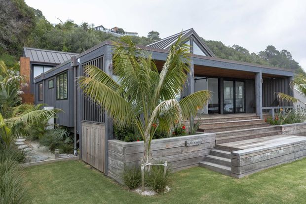 Exterior view of a compact contemporary home set into a lush, sloping coastal landscape. The building features dark grey vertical cladding, standing-seam metal gable roofs, large glazed openings, timber soffits, and broad timber-clad entry steps and raised garden beds. Tropical palms and dense planting surround the front terrace.