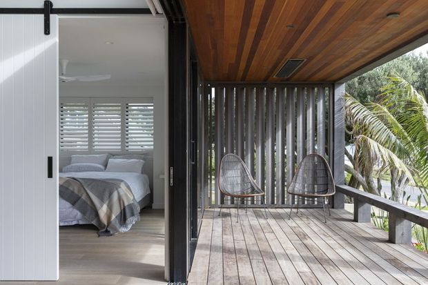 A timber-clad covered balcony or terrace adjoining a bedroom through an open white sliding barn-style door. The outdoor deck has weathered timber floorboards, dark vertical privacy screens, a timber-lined ceiling, two woven lounge chairs, and a dark timber or metal balustrade. The bedroom visible inside has pale finishes, plantation shutters, and neutral bedding. Lush tropical vegetation is visible beyond the terrace.