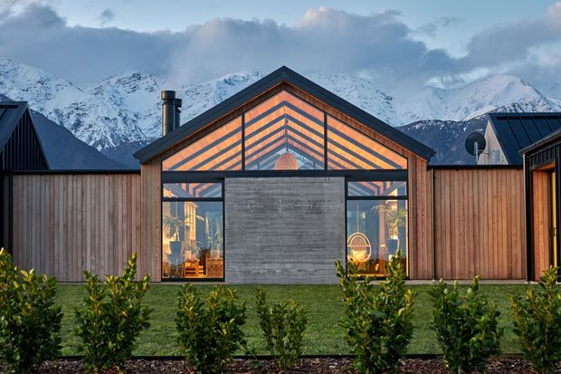 A compact modern gabled house or pavilion set against dramatic snow-covered mountains. The facade combines vertical timber fencing and cladding with dark metal roof framing, extensive glazed gable and windows, and a central grey concrete or masonry feature wall. Warm interior lighting and visible planting create contrast with the cool alpine setting.