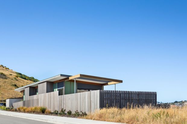 Exterior view of a low, contemporary single-storey house on a sloping site. The building has a flat, layered roofline with broad eaves, extensive glazing, vertical timber screening and fencing, and a muted grey-brown facade palette. Dry grasses and a hillside surround the property under a clear blue sky.