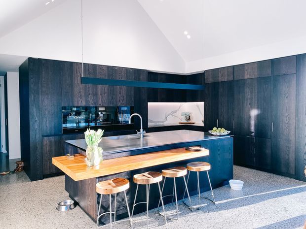 A contemporary kitchen with dark timber cabinetry spanning the walls, a large blue-toned island, stainless-steel benchtop, timber breakfast bar, and four timber stools. A marble-look splashback, integrated appliances, pendant-style linear lighting, and a high vaulted white ceiling complete the bright, refined space.