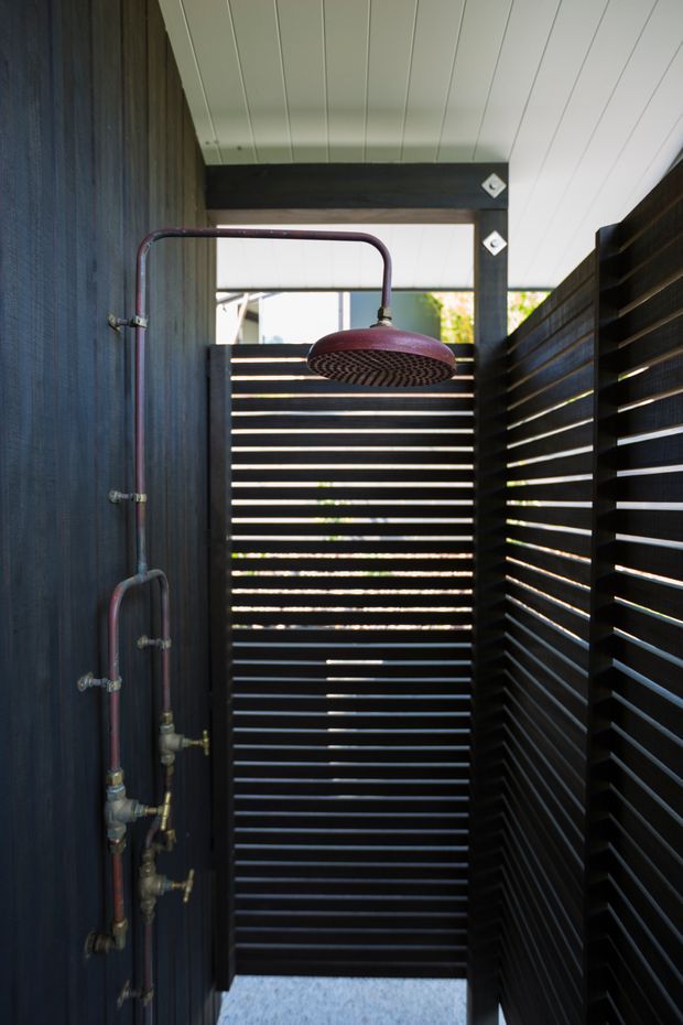 A compact outdoor shower enclosure with exposed copper plumbing, a large round showerhead, dark horizontal slatted privacy screens, and a white-lined ceiling. The space is narrow, partially open to daylight, and finished in dark timber-like materials with metallic plumbing details.