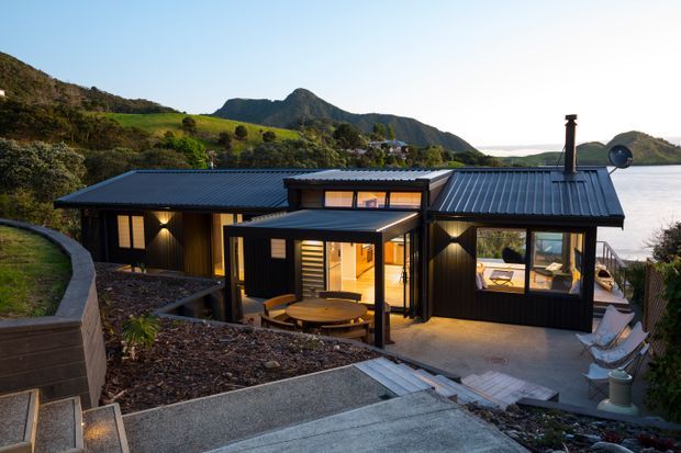 A modern single-storey dark-clad house set into a steep landscaped hillside, with multiple pitched metal roof forms, extensive glazing, and warm interior lighting. A paved and stepped approach leads to a covered outdoor dining area and sheltered terrace. Rugged green hills and a body of water are visible behind the house.