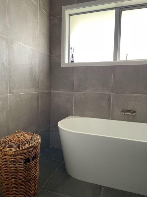 A contemporary bathroom featuring a freestanding white bathtub, large dark grey wall and floor tiles, a white-framed frosted window, and a woven wicker basket. The space has a clean, understated design with natural texture from the basket.