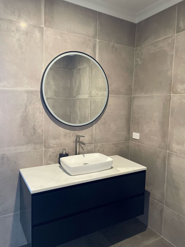 A contemporary bathroom vanity with a white vessel basin, dark blue-black floating cabinetry, pale stone benchtop, brushed metal tapware, and a large round framed mirror. The walls and floor are finished in large-format grey-beige stone-look tiles, creating a clean, restrained palette.