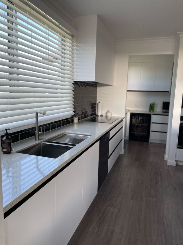 A bright, contemporary galley kitchen with white flat-panel cabinetry, dark contrasting trim, a pale speckled stone benchtop, and a dark glossy tiled splashback. The kitchen includes a double stainless-steel sink beneath wide horizontal window blinds, integrated appliances, and dark timber-look flooring. A compact pantry or secondary kitchen area is visible at the rear.