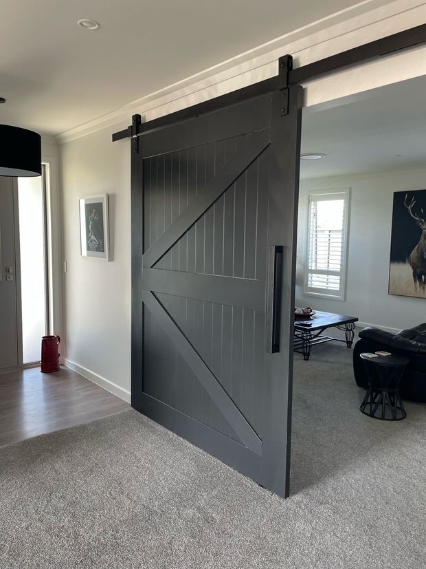 Interior view of a residential hallway and living room separated by a large dark charcoal barn-style sliding door on exposed black metal hardware. The door features vertical board panelling and diagonal bracing. The hallway has pale walls, timber-look flooring and a black pendant light, while the adjoining living room has grey carpet, a black leather sofa, coffee table and a window with white shutters.