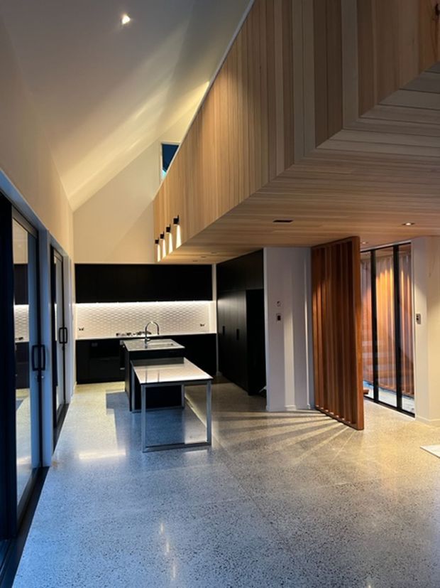 Open-plan contemporary kitchen and living area with polished terrazzo flooring, black cabinetry, a central island, and a light geometric tile splashback. Warm timber wall and ceiling panelling, a dramatic sloping ceiling, extensive dark-framed glazing, and a large vertical timber pivot screen create a refined, warm modern interior.