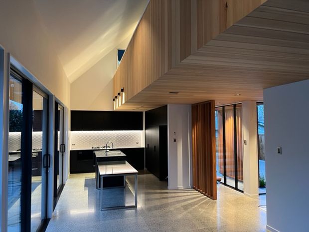 Contemporary open-plan kitchen and living area with a high sloping ceiling, extensive glazing, black cabinetry, a central island, and a warm timber-lined ceiling and joinery feature. The polished light floor and layered lighting create a refined, spacious interior.