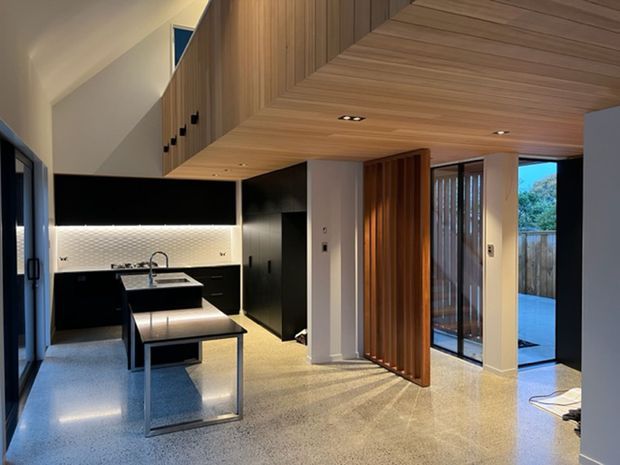 A contemporary open-plan kitchen and living area with black cabinetry, a compact island with an integrated sink, and a textured light backsplash. The space features a warm timber-lined ceiling continuing into overhead joinery, polished terrazzo flooring, dark-framed glazing, and a timber-screened opening leading to an outdoor deck or courtyard. Recessed lighting and concealed illumination create a refined, high-contrast interior.
