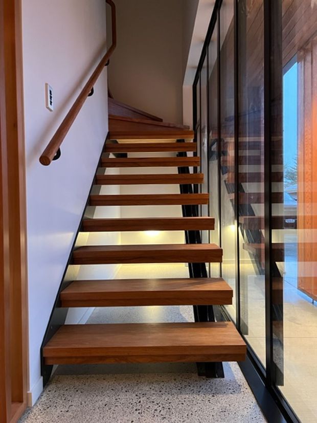 Interior view of a contemporary open-riser staircase with warm timber treads, slim dark metal supports, and a timber handrail. The stair runs beside full-height black-framed glazing, with polished terrazzo flooring and reflected interior surfaces visible beyond the glass.