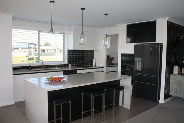 Contemporary kitchen with a large light-coloured island and dark base cabinetry, black tiled splashback, integrated black refrigerator, and three pendant lights. White cabinetry and plantation shutters surround a bright window, while a television and dark feature wall are visible at the rear.