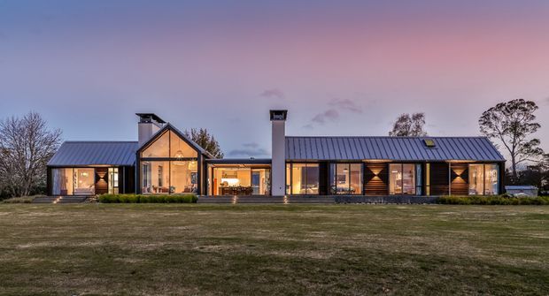 Wide dusk exterior view of a long, low-slung contemporary single-storey residence set in an open landscaped lawn. The house has dark metal pitched roofs, extensive warm-lit glazing, timber-clad wall sections, a prominent glazed gabled pavilion, and multiple chimney forms. Mature trees and a pastel evening sky form the background.