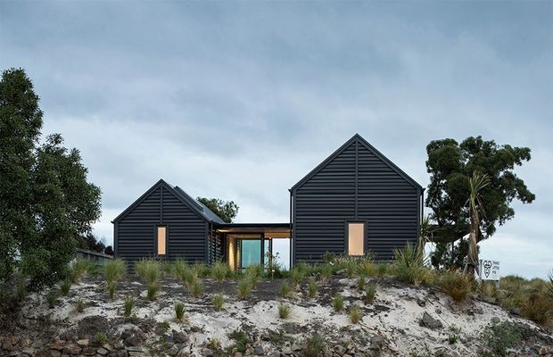 Exterior view of a contemporary coastal-style residence composed of two dark horizontal-clad gabled volumes connected by a glazed central link. The house sits on a sandy, sparsely landscaped site with native grasses and coastal vegetation under an overcast sky. Vertical timber-toned entry or window elements punctuate the dark facades.