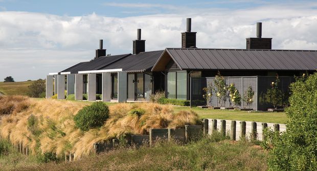 A low, elongated contemporary residential building set in an open rural or coastal landscape. The dark standing-seam pitched roof and dark cladding contrast with pale vertical wall panels and extensive glazing. Multiple chimney flues rise from the roof, while grasses, shrubs, and a low timber retaining structure occupy the foreground.