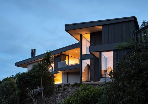 Twilight exterior view of a contemporary multi-level residence set on a landscaped sloping site. The home features dark vertical cladding, crisp flat roof planes, extensive floor-to-ceiling glazing, projecting upper volumes, balconies, and warm interior lighting visible through the windows. Dense planting and rocky ground frame the facade.