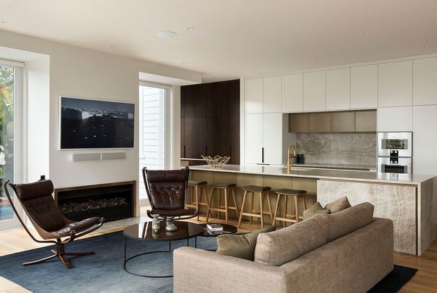 Contemporary open-plan living room and kitchen with a neutral palette, built-in white cabinetry, dark timber joinery, stone-look island and splashback, timber flooring, and a fireplace. The living area includes a large sofa, leather lounge chairs, coffee tables, and wall-mounted television, with bar seating at the kitchen island.