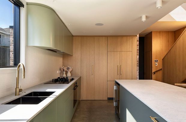 A compact contemporary kitchen with full-height light timber cabinetry, muted sage-green lower cabinets, a pale stone benchtop, integrated stainless appliances, and a large central island. A curved rangehood enclosure and curved upper cabinetry soften the linear joinery. The space includes a tiled splashback, dark polished concrete-like flooring, black sink, brushed brass tapware, and a warm timber-lined entry or adjoining area.