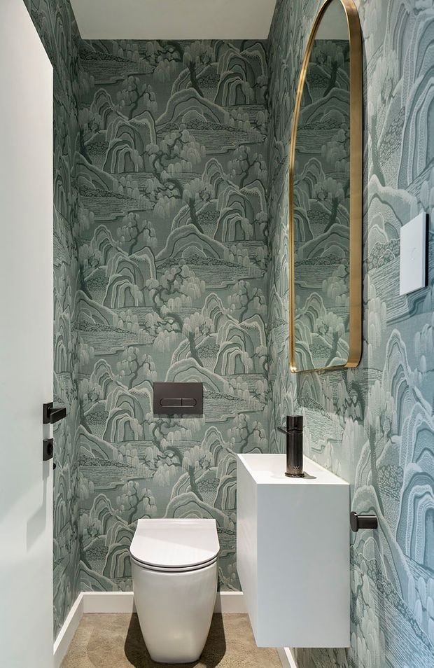 A compact powder room with a wall-mounted white toilet and a minimalist white cube vanity with a black tap. The walls are covered in distinctive muted green and grey scenic patterned wallpaper, complemented by a large vertically oriented arched mirror with a brass-toned frame. Dark hardware and a concealed cistern plate provide contrast, while the floor is finished in a neutral textured material.