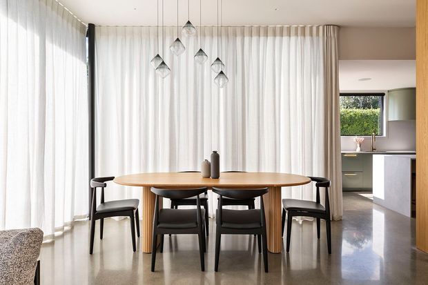 Bright contemporary dining area with a large round timber dining table and six dark chairs, set against full-height sheer curtains and polished concrete flooring. A cluster of pendant lights hangs above the table, while an open connection to the kitchen is visible at right.