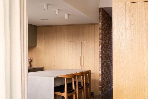 A warm, contemporary kitchen viewed through a doorway, featuring a large pale stone island with timber bar stools, extensive light timber floor-to-ceiling cabinetry, integrated handles, a dark benchtop or side surface, and an exposed red-brown brick wall. The ceiling has simple recessed and surface-mounted downlights, with a soft neutral palette and clean joinery.