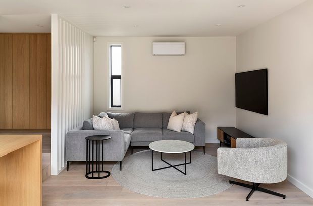 A bright, compact contemporary living room viewed from an adjoining kitchen. The room features a grey sectional sofa, textured light armchair, round coffee table, black nesting side table, wall-mounted television, split-system air conditioner, pale timber flooring, and a white vertical slatted divider. A narrow black-framed window brings in natural light, while pale neutral walls and furnishings create a clean, understated interior.
