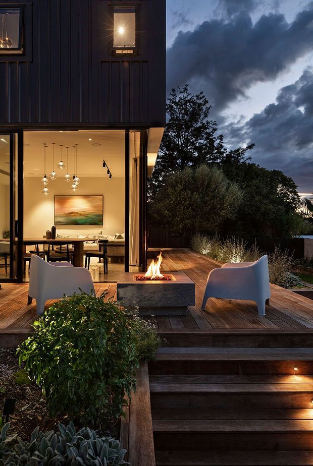 A contemporary multi-level home at dusk, viewed from a landscaped timber deck and stairway. Large black-framed glazed doors open onto an illuminated dining and living area, while the exterior deck features two lounge chairs, a central outdoor fire pit, built-in lighting, and dark timber cladding. Dense planting and dramatic evening clouds create a warm indoor-outdoor setting.