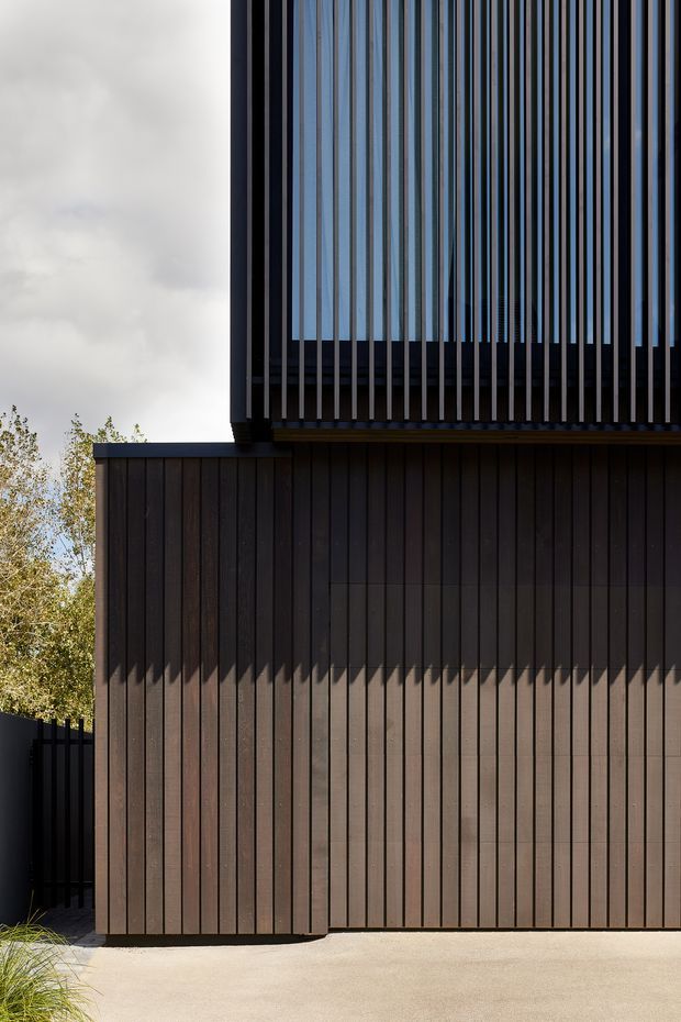Close-up exterior view of a contemporary multi-level building with dark vertical timber cladding, a projecting upper volume, and a tall screened facade of closely spaced vertical fins. A pale paved surface and landscaping sit at the base, with soft daylight and tree foliage in the background.