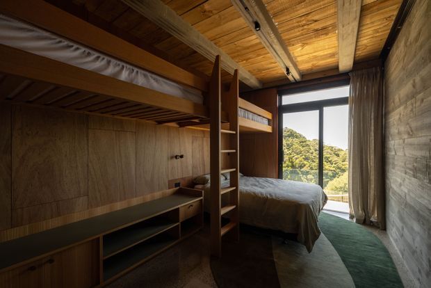 Compact, warm-toned bunk bedroom with custom timber joinery. A raised bunk occupies the left side above built-in shelving, desk and storage, while a lower double bed sits beside a full-height glazed door or window opening to leafy views. The room has exposed timber ceiling boards and beams, wood wall panelling, a textured masonry side wall, neutral bedding, and a muted green runner. Natural light creates a cosy, cabin-like atmosphere.