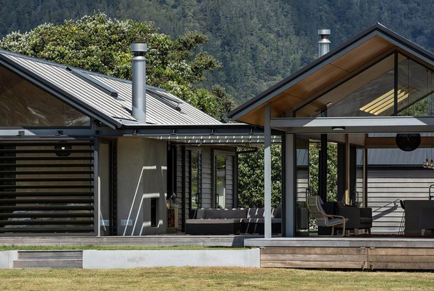Exterior view of a contemporary single-storey home set against a wooded mountain backdrop. The building has multiple pitched gabled roof forms with dark metal roofing, exposed timber soffits, extensive glazing, grey rendered walls, a covered timber deck, and a prominent metal flue. A louvred screen and outdoor seating are visible beneath the sheltered areas.