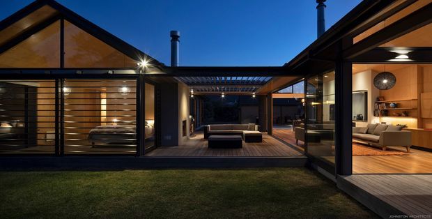 Dusk view of a modern single-storey courtyard-style residence arranged around a central outdoor living space. The composition features extensive floor-to-ceiling glazing, dark structural frames, warm timber-lined interiors, timber decking, a covered central terrace, and landscaped lawn in the foreground. Living areas and furnishings are visible through the glazing.