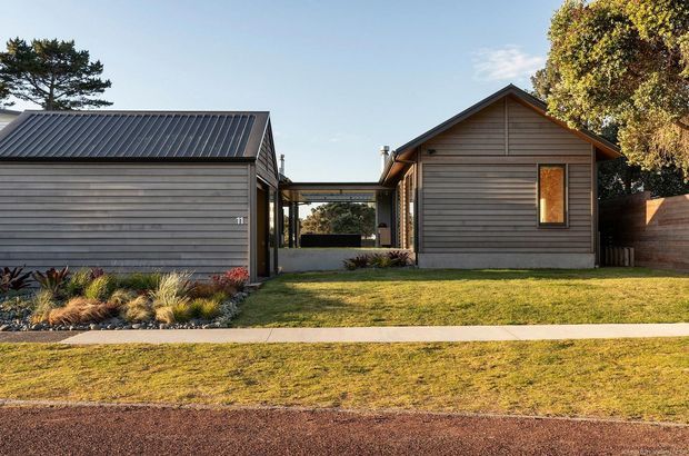Exterior view of a low-rise residential property composed of two timber-clad gabled volumes linked by a glazed or open central passage. The left volume has a dark corrugated metal roof and garage-like frontage, while the right volume has horizontal weatherboard cladding, a pitched gable roof, and a narrow window. A neat front lawn, simple planting beds, paved footpath, and timber boundary fence complete the suburban streetscape.