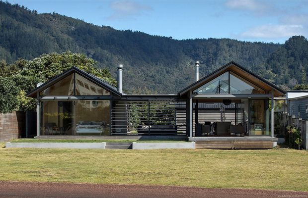 Single-storey contemporary residential building set against a forested mountain backdrop. The facade consists of two glazed gabled volumes linked by a central dark horizontal slatted screen and covered outdoor connection. Dark metal roof framing, timber-toned soffits, extensive glazing, chimneys, and a broad lawn are visible.