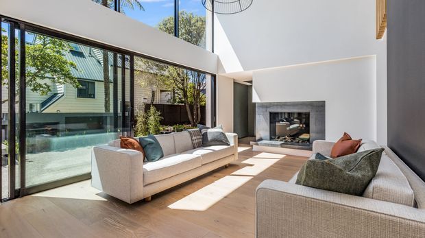 A bright contemporary living room with a double-height ceiling, expansive floor-to-ceiling glazed sliding doors, pale timber flooring, and two large light upholstered sofas arranged around a recessed fireplace with a dark stone surround. Strong sunlight enters from the glazing, with views to a landscaped courtyard and neighbouring architecture outside.