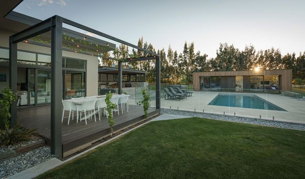 Contemporary residential outdoor entertaining area with a covered pergola and outdoor dining setting beside a modern home. The space opens onto a paved pool terrace with a rectangular swimming pool, lounge chairs, lawn, planted borders, and a separate low-profile pool pavilion or ancillary structure. Dark metal framing, timber decking, large glazed doors, and horizontal timber-clad elements define the scene.