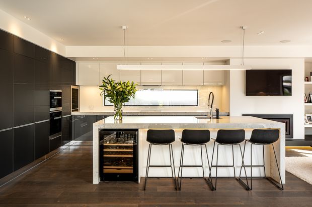 Contemporary open-plan kitchen with a large white island and four black bar stools, dark full-height cabinetry, integrated appliances, a wine fridge, pale overhead cabinetry, timber flooring, stone benchtops, and linear pendant lighting. A living area with wall-mounted television is visible beyond the kitchen.