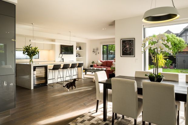 Bright open-plan residential interior combining a contemporary kitchen, dining area and living room. The kitchen features a large island with waterfall stone benchtop, dark cabinetry, integrated appliances and bar seating. A dining table with upholstered chairs sits beside large windows overlooking a landscaped garden, while a lounge area with a red sofa is visible beyond. Dark timber flooring, white walls, pendant lighting and indoor plants create a warm, refined atmosphere.