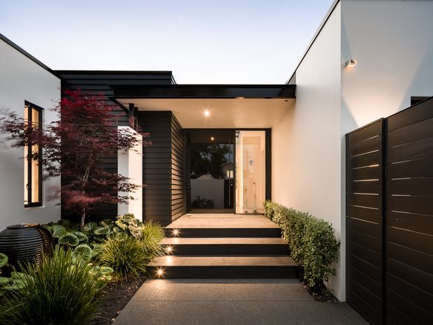 A contemporary residential entry viewed from the front path, featuring crisp white rendered walls, dark horizontal cladding, a flat projecting canopy, illuminated steps, a glazed front door, and a landscaped garden bed with ornamental planting. A tall dark privacy gate lines the right side.