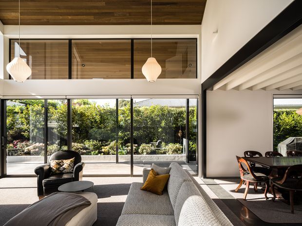 Bright open-plan living room with a large textured sectional sofa, black leather armchair, and low coffee table. Floor-to-ceiling glazing overlooks a landscaped courtyard, while a dining area is visible to the right. The room features exposed dark timber ceiling lining, black-framed internal glazing, white walls, and distinctive pendant lights, with strong indoor-outdoor connection and abundant natural light.