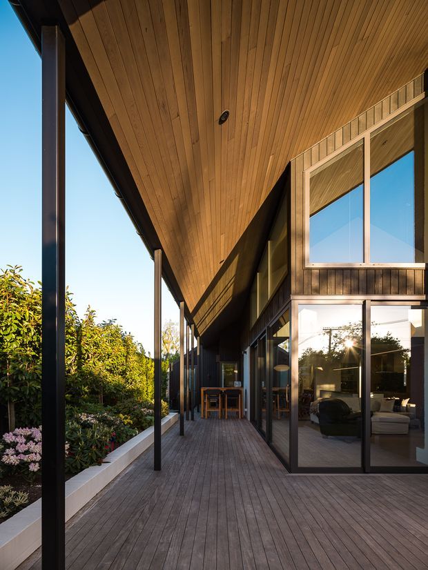 A covered timber deck and veranda runs alongside a contemporary home, with a warm timber-lined soffit, dark slender structural posts, timber decking, and full-height glazed doors and windows. The glazing reveals a living room and dining area, while landscaped planting borders the deck. Strong linear perspective and abundant natural light define the image.