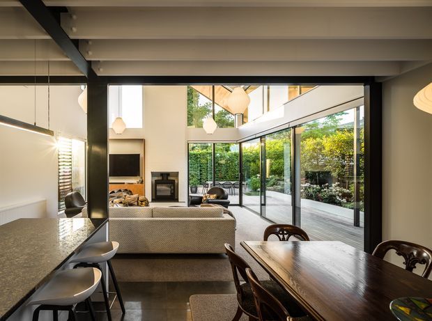 Open-plan contemporary living, dining, and kitchen space with exposed dark structural beams, a polished stone kitchen island, timber dining table, upholstered seating, a fireplace, and extensive floor-to-ceiling glazing opening onto a leafy courtyard or garden. The room has a bright, airy feel with strong indoor-outdoor connection.