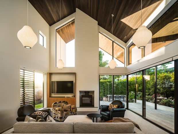 A spacious contemporary living room with a dramatic double-height ceiling, dark timber ceiling lining, exposed roof geometry and extensive floor-to-ceiling glazing. The room features a large sectional sofa, leather lounge chairs, patterned cushions, a built-in television niche, fireplace, sculptural pendant lights and views into a landscaped outdoor area.