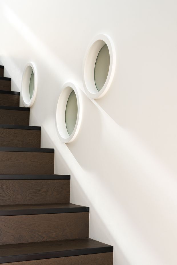 A minimalist stairway with dark timber stair treads and risers, contrasted by black-edged nosings and a smooth white wall. Three oval recessed wall features with pale translucent centers add sculptural detail and cast soft diagonal shadows across the wall. The composition is bright, clean, and contemporary.