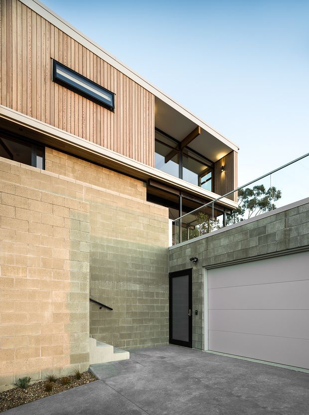 Contemporary multi-level residential exterior viewed from a driveway or courtyard, featuring a warm vertical timber-clad upper volume above exposed concrete block walls. The composition includes a recessed glazed balcony with a clear glass balustrade, a narrow horizontal window, dark-framed doors and windows, an integrated white sectional garage door, concrete paving, and restrained exterior lighting. The building has a strong modern geometric form with contrasting natural and masonry materials.