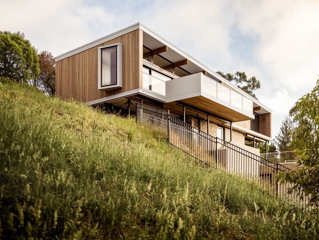 Exterior view of a contemporary multi-level residence built into a steeply sloping, landscaped site. The house features warm vertical timber cladding, crisp white balcony planes and trim, extensive glazing, projecting balconies, and dark metal balustrades. The elevated form and stepped massing follow the hillside.