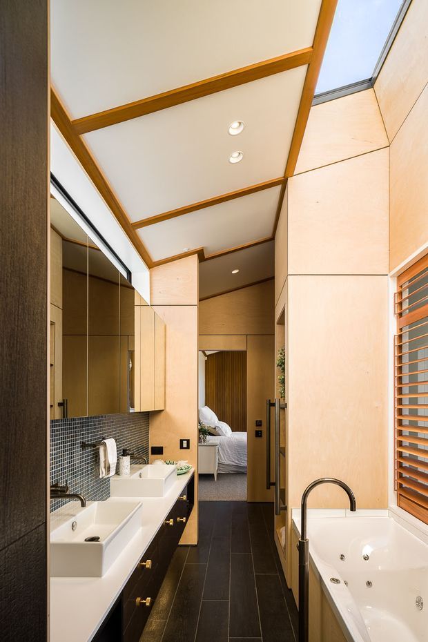 A narrow, refined bathroom with a double vanity and freestanding spa bath. The space features pale timber wall panelling and cabinetry, dark tiled flooring, a small dark mosaic splashback, mirrored storage, brass-toned hardware, timber ceiling beams, recessed downlights, and a skylight. A bedroom is visibly connected beyond the bathroom, while timber louvred glazing brings in daylight.