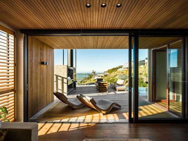 A warm contemporary interior opening onto a timber deck and coastal outlook. The room features extensive black-framed sliding glass doors, a richly timber-lined ceiling and wall, timber flooring, and built-in window shutters. Two sculptural lounge chairs face the outdoor terrace, where additional seating and a built-in outdoor kitchen or barbecue are visible. Sunlight creates strong linear shadows across the floor, with ocean and hillside views beyond.
