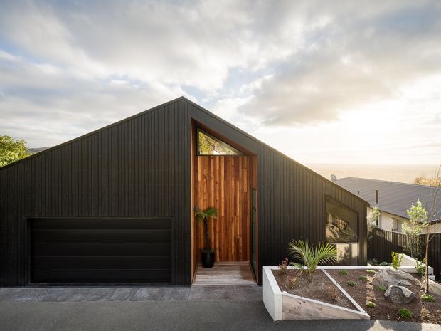 Contemporary dark-clad gabled house facade viewed from the street, featuring a recessed timber entry door beneath a triangular glazed opening, an integrated black garage door, and a raised landscaped front garden with retaining walls. The composition is minimalist and strongly geometric, with warm timber contrasting against charcoal vertical cladding.